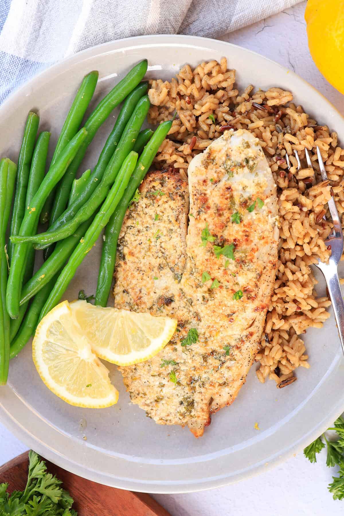 A plate with a broiled Parmesan tilapia filet, steam green beans, brown rice, and lemon wedges.