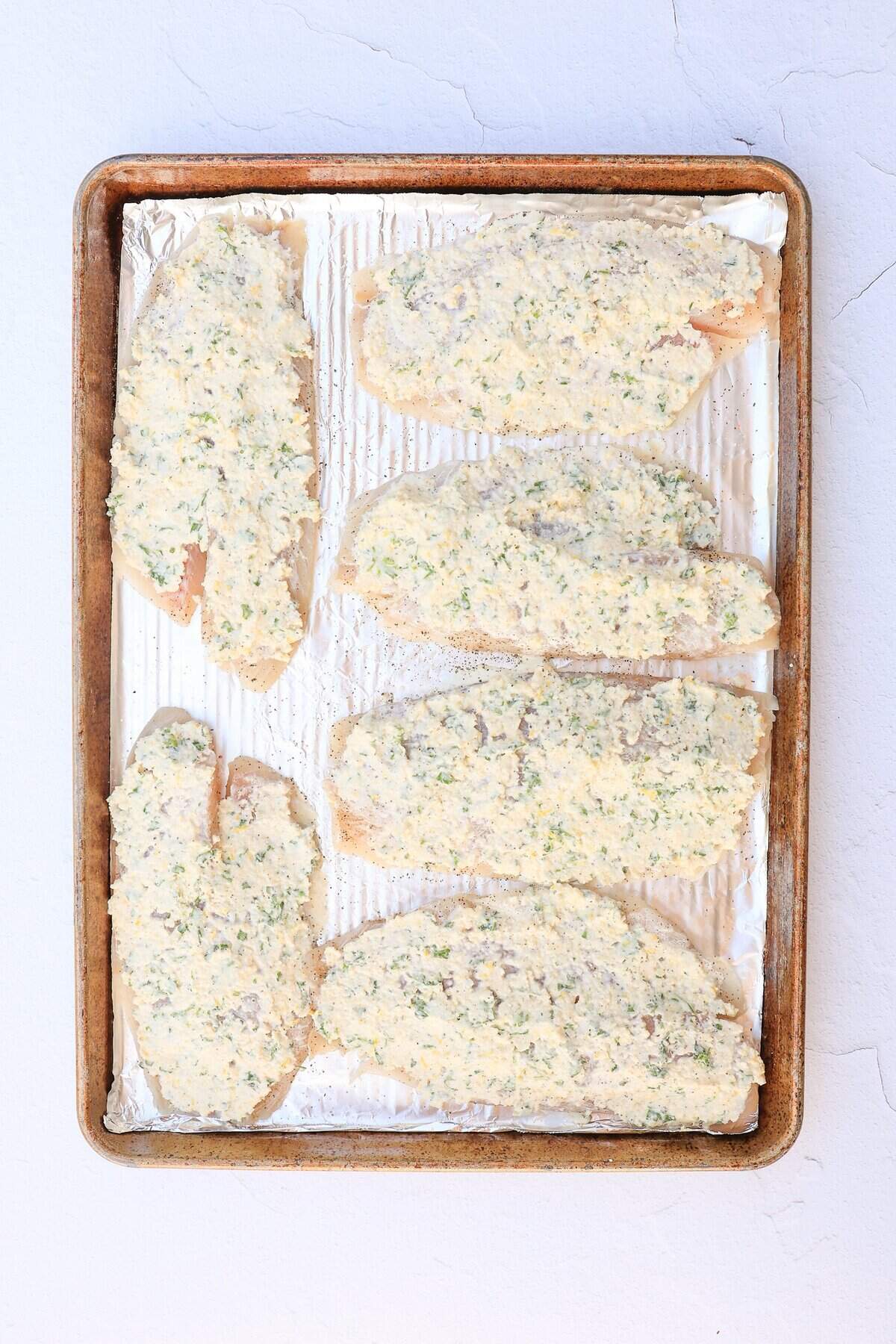 Tilapia filets on a foil-covered baking sheet smothered in a mixture of Parmesan, butter, mayonnaise, and seasonings before being broiled.