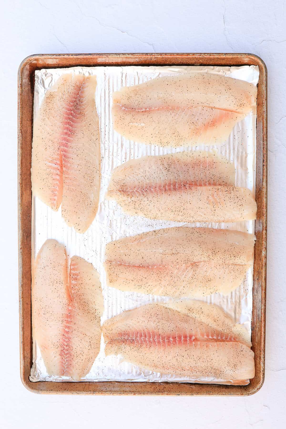 Raw tilapia filets on a foil-covered baking sheet seasoned with salt & pepper.