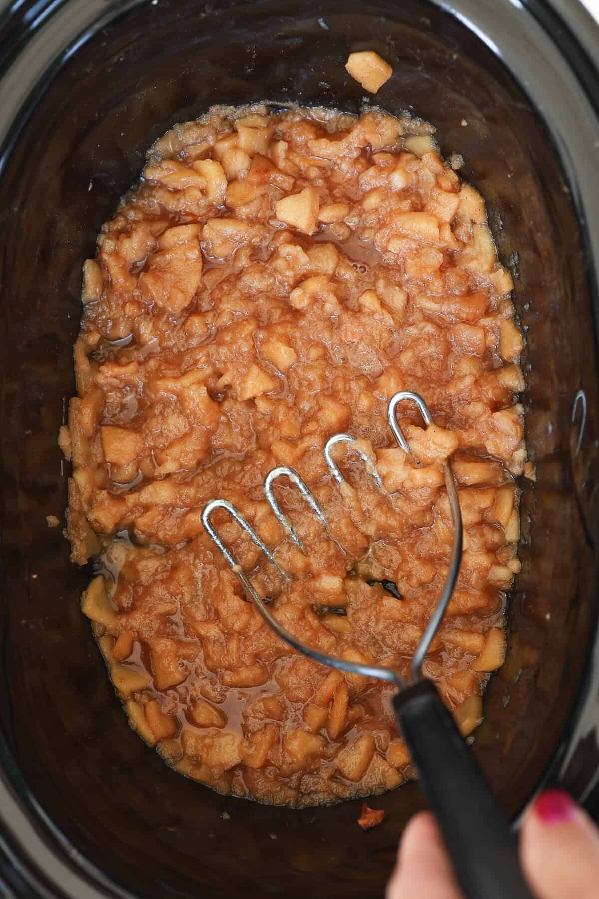 A potato masher smashing cooked apples in a crockpot.