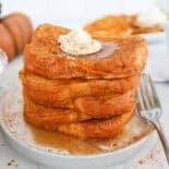 Thick pumpkin french toast slices stacked on a plate with maple syrup, whipped butter, and cinnamon on top.