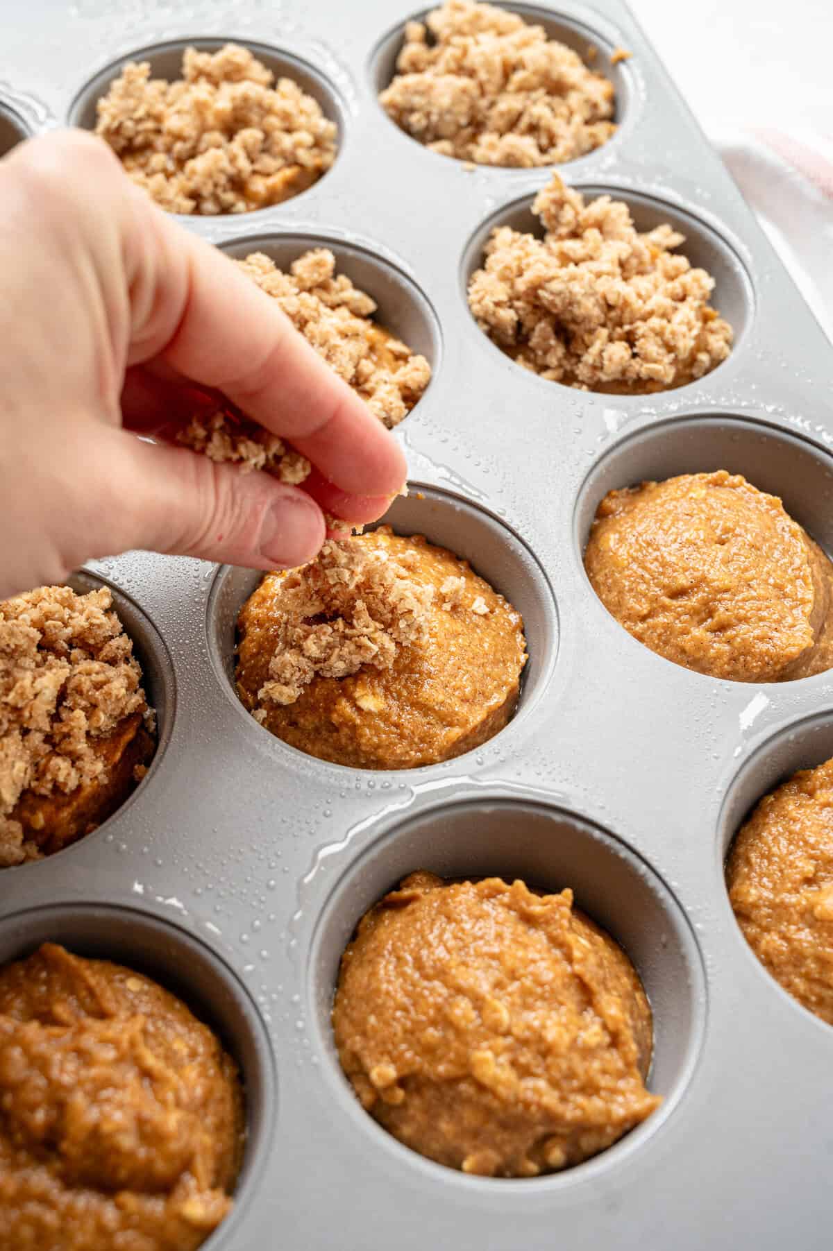 Crumble topping being sprinkled on top of pumpkin batter in a muffin tin.