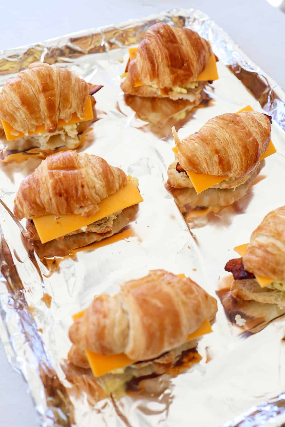 Croissant breakfast sandwiches on a foil-lined baking sheet ready for the oven.
