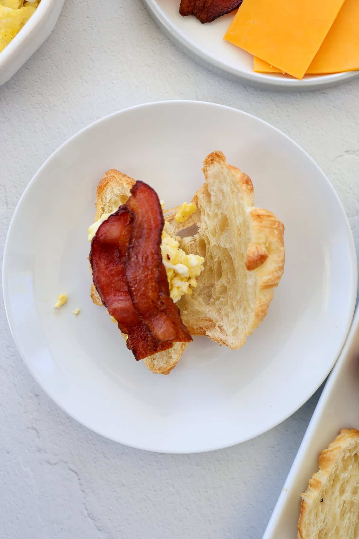 A croissant split in half with scrambled eggs and cooked bacon on it.
