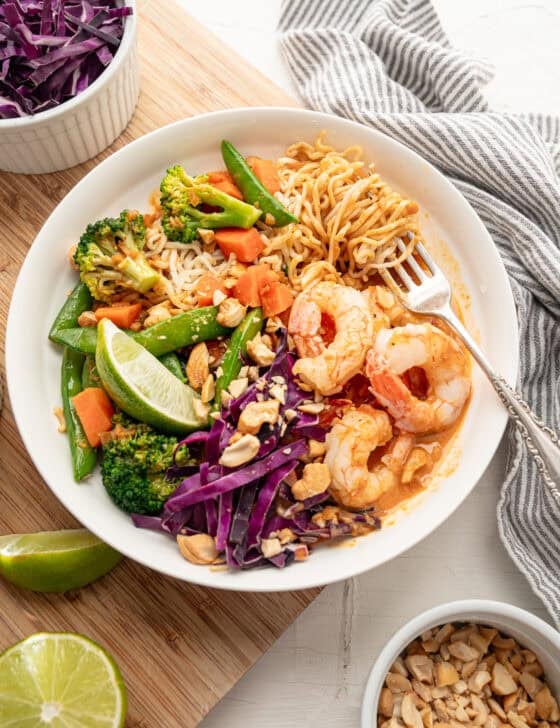 A shrimp curry bowl with bowls of chopped peanuts, lime wedges, and shredded cabbage on the side.
