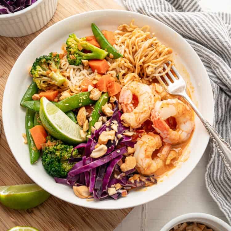 A shrimp curry bowl with bowls of chopped peanuts, lime wedges, and shredded cabbage on the side.