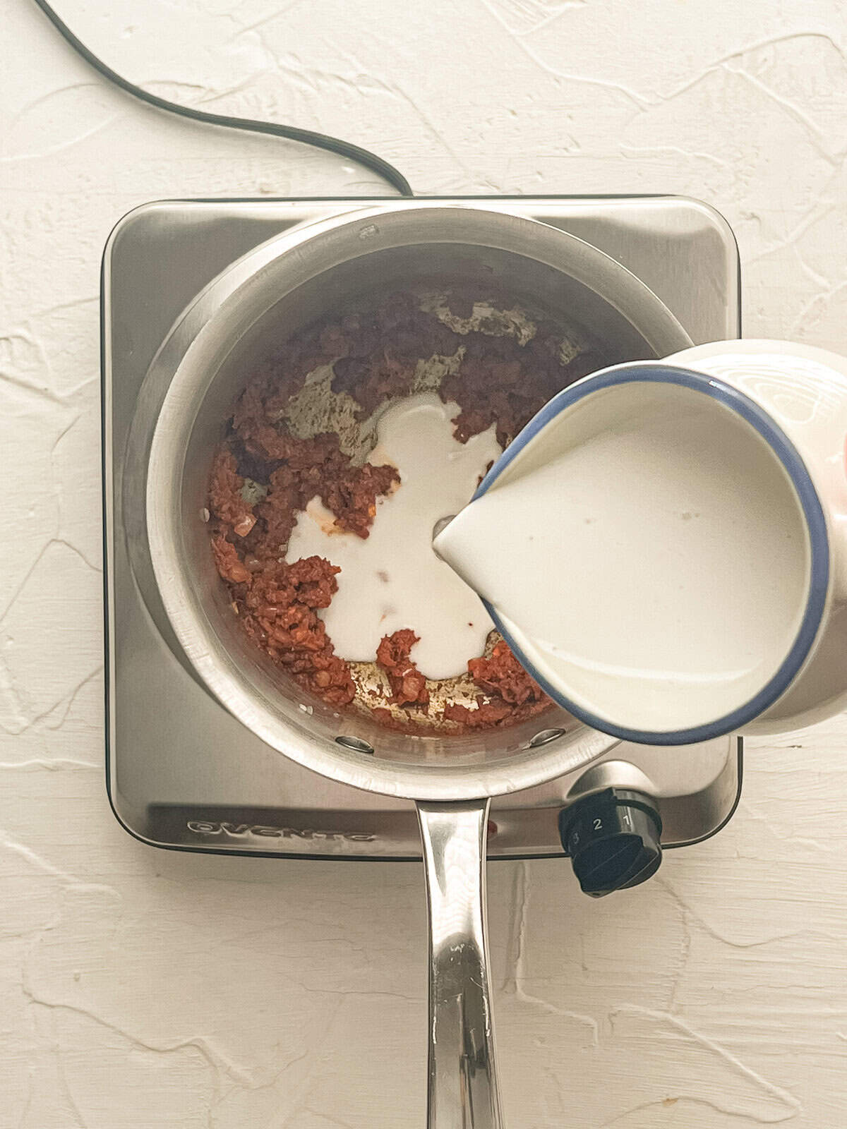 Coconut milk being poured into a sauce pan with curry paste and sauteed onions.