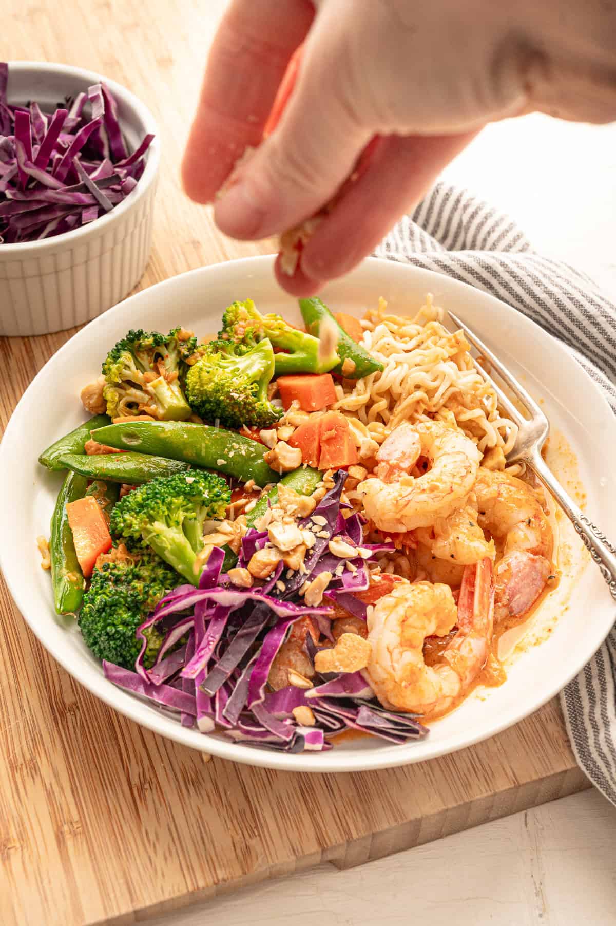 A hand sprinkling chopped peanuts on top of a shrimp curry bowl.
