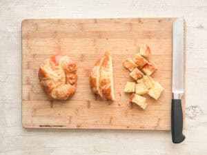 Croissants on a wooden cutting board being chopped in to cubes.