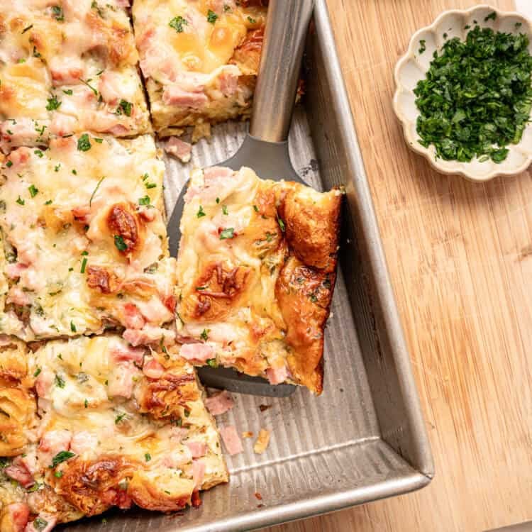 A serving of croissant breakfast casserole being lifted out of a baking dish.