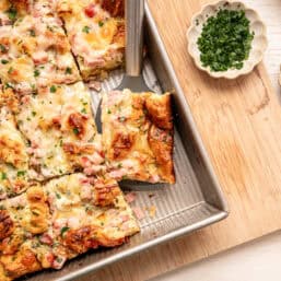 Croissant breakfast casserole being served from a baking dish with fresh herbs on top.
