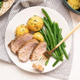 Sliced pork tenderloin on a plate with boiled baby potatoes and green beans.