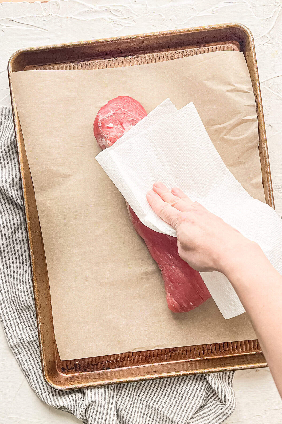 A hand patting a raw pork tenderloin with paper towels.