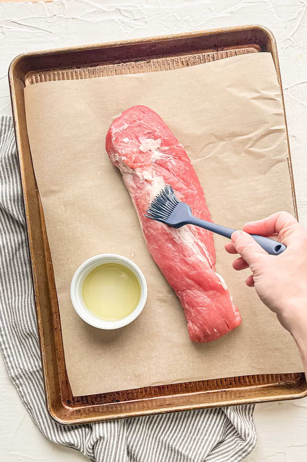 A hand brushing oil on a pork tenderloin on a baking sheet.