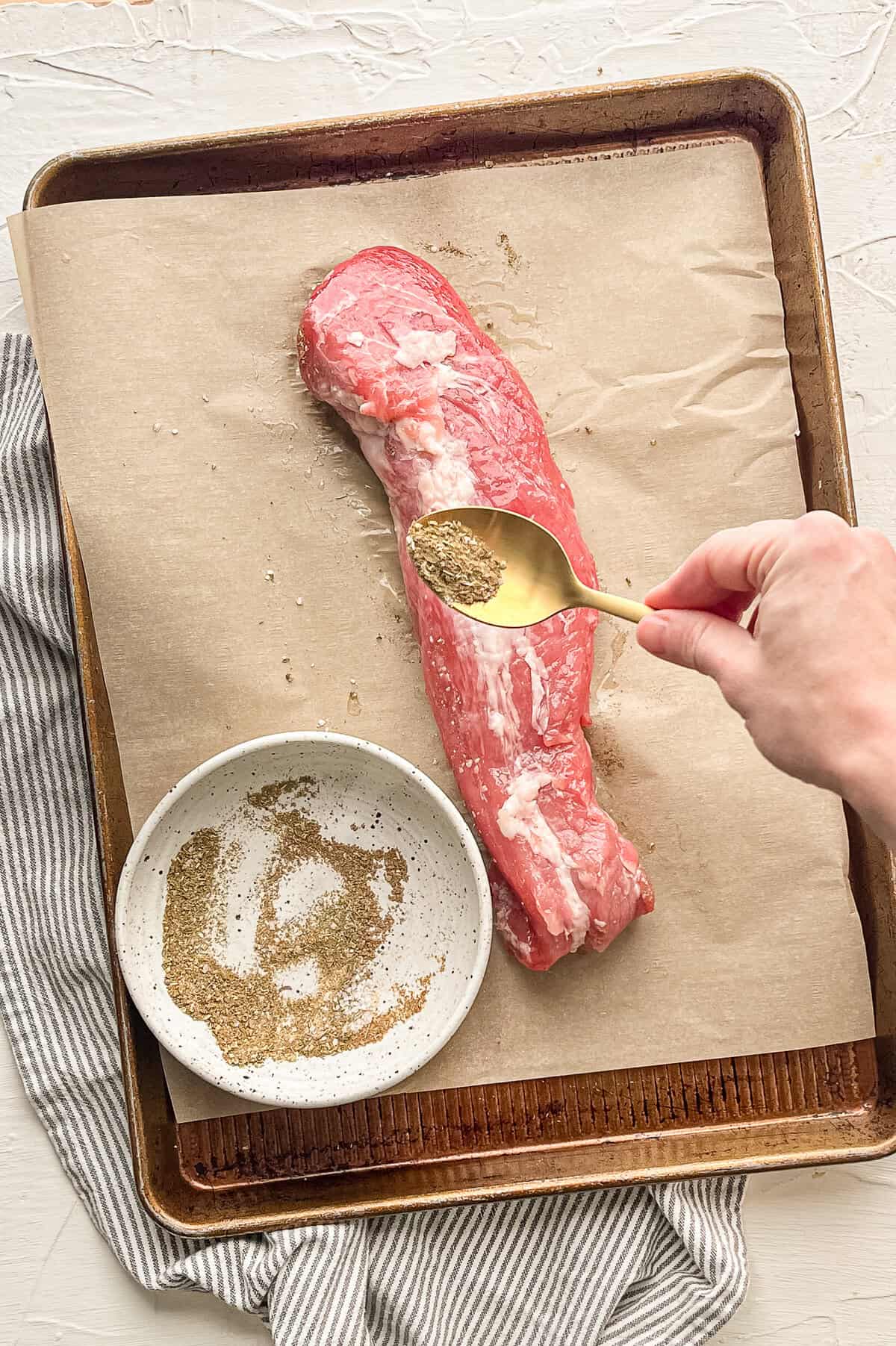 A hand sprinkling a mix of ground seasonings being put on a pork tenderloin before roasting.