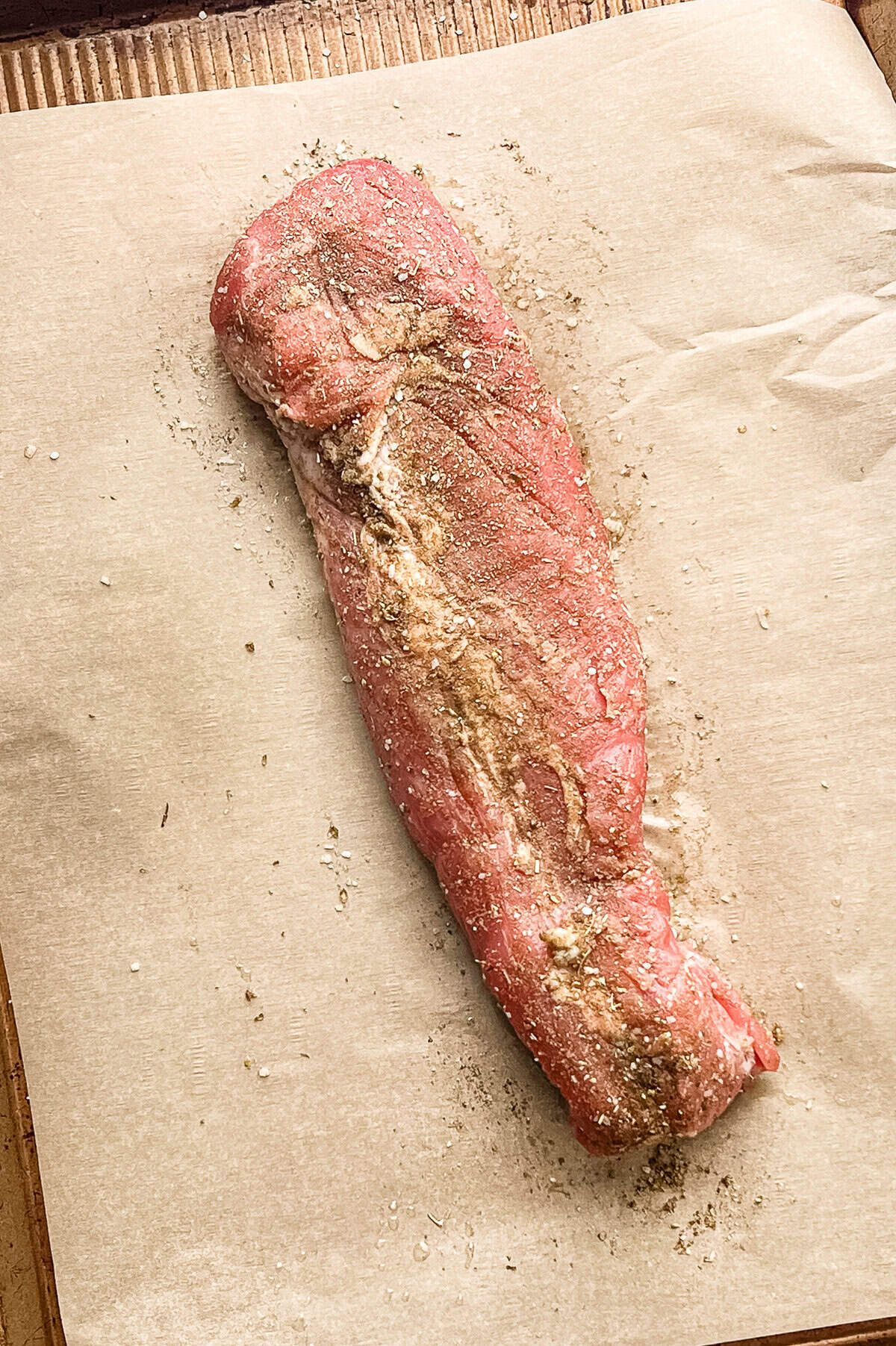 Pork tenderloin on a parchment-lined baking sheet with a mix of dried seasonings on it.