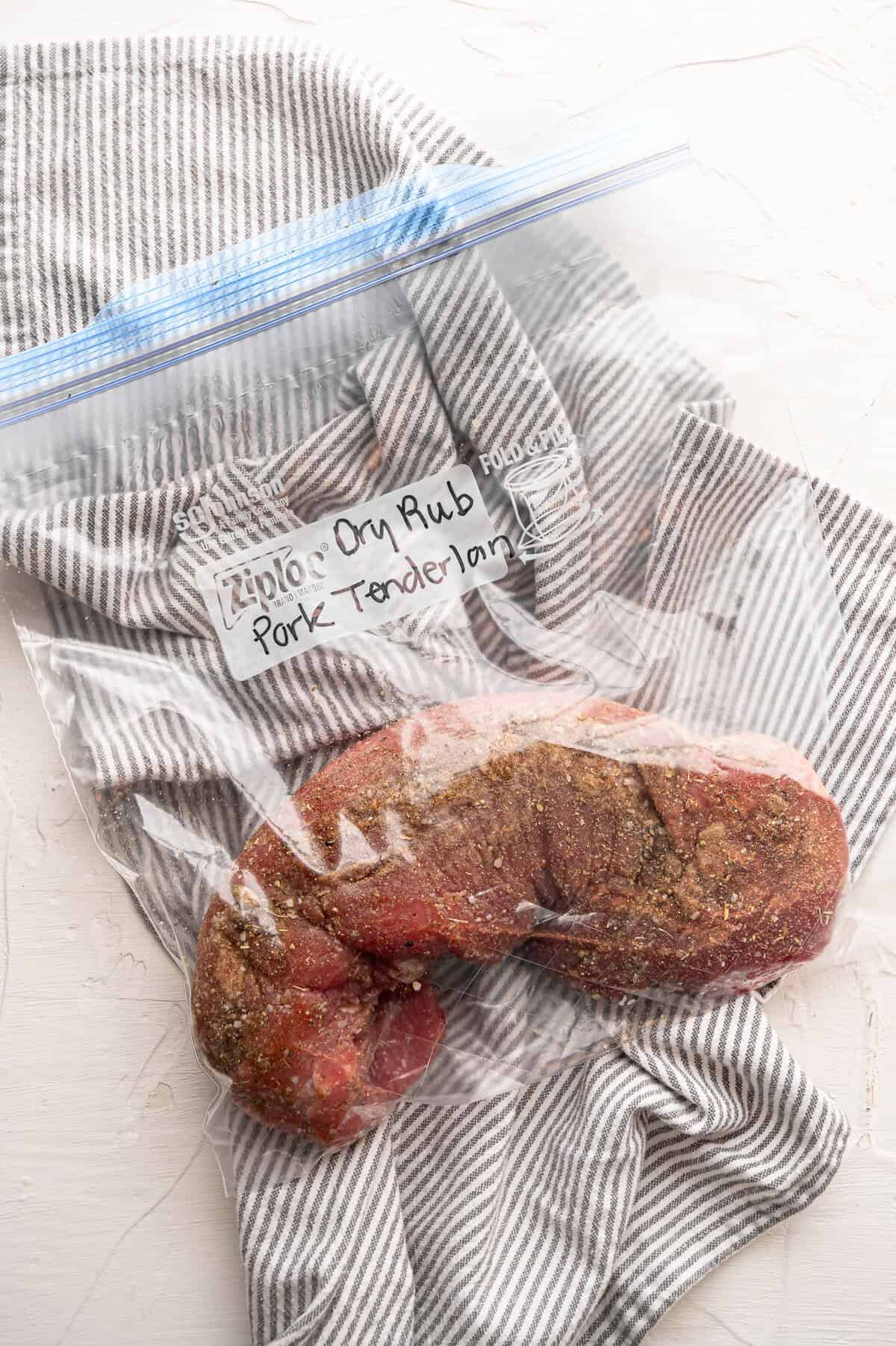 A raw pork tenderloin rubbed with ground seasonings inside a freezer bag.