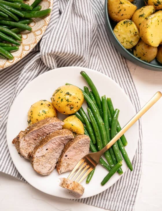 Sliced pork tenderloin on a plate with boiled baby potatoes and green beans with a small bite of pork on a fork.