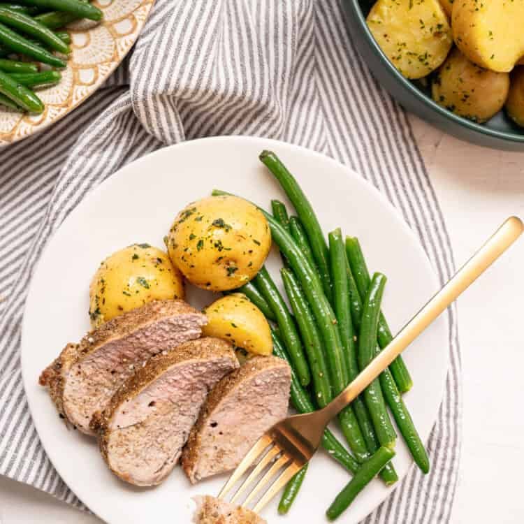 Sliced pork tenderloin on a plate with boiled baby potatoes and green beans with a small bite of pork on a fork.