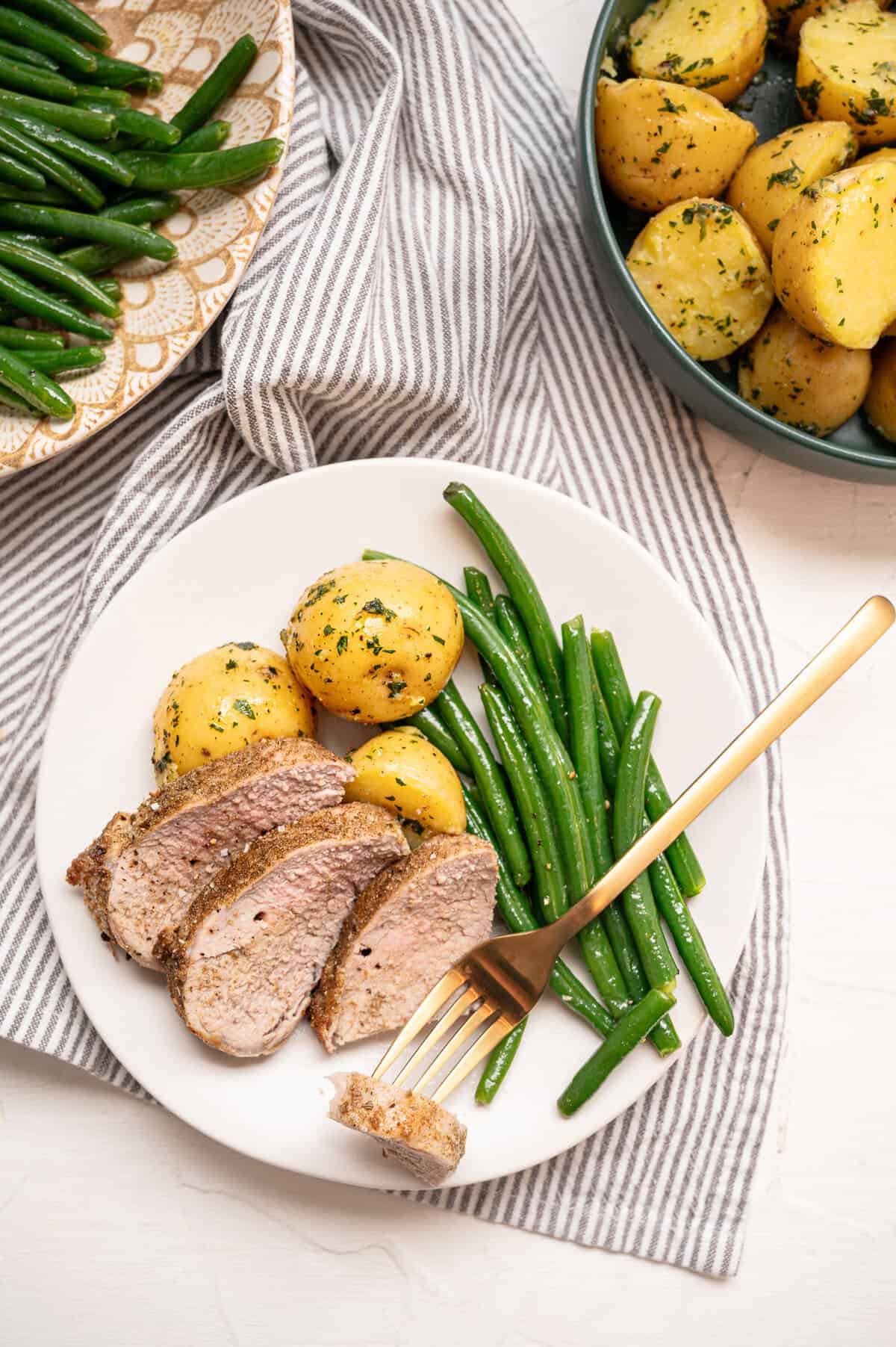 Sliced pork tenderloin on a plate with boiled baby potatoes and green beans with a small bite of pork on a fork.