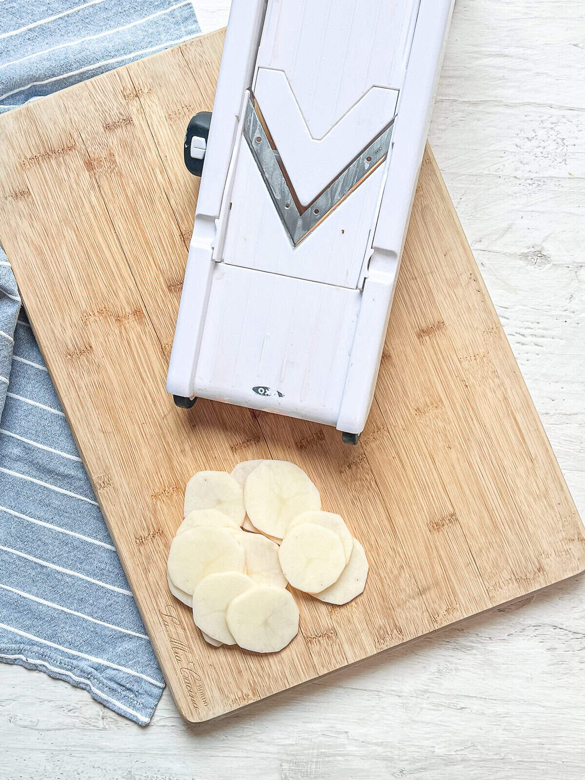 Thinly sliced Russet potatoes on a cutting board next to the mandolin that was used to slice them.