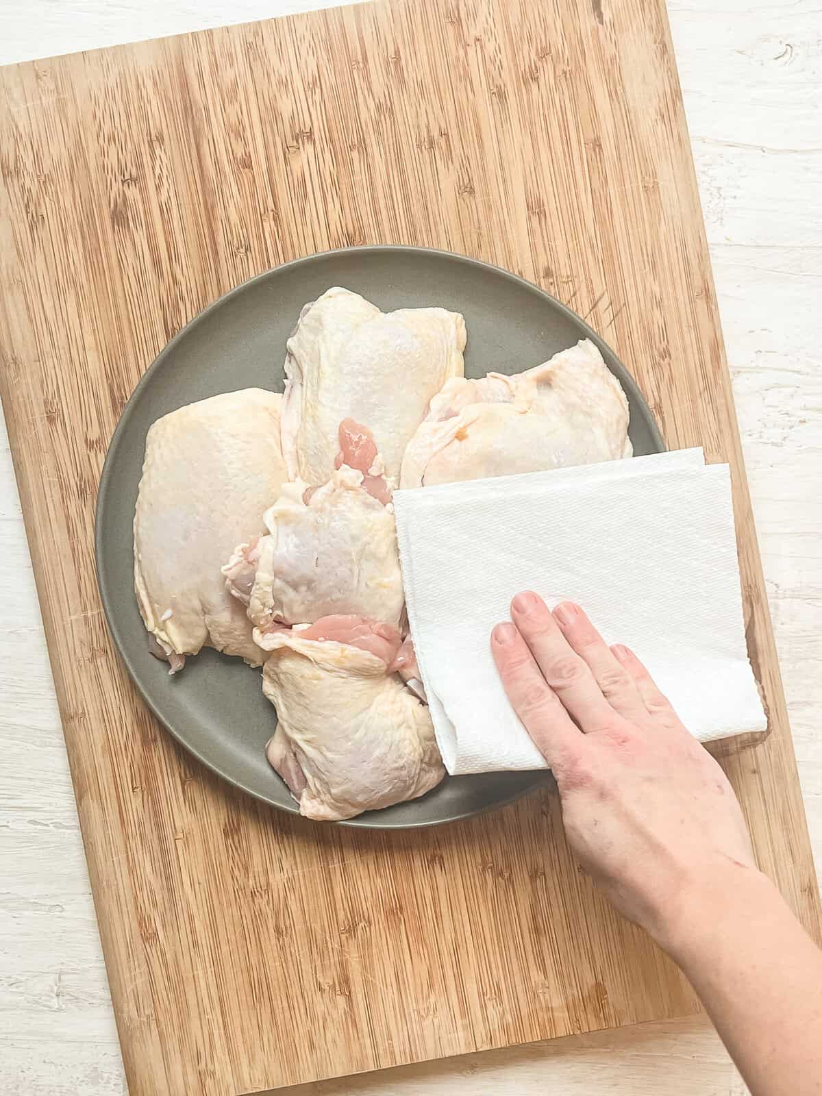 A hand dabbing paper towels on bone-in, skin-on chicken thighs to remove moisture.