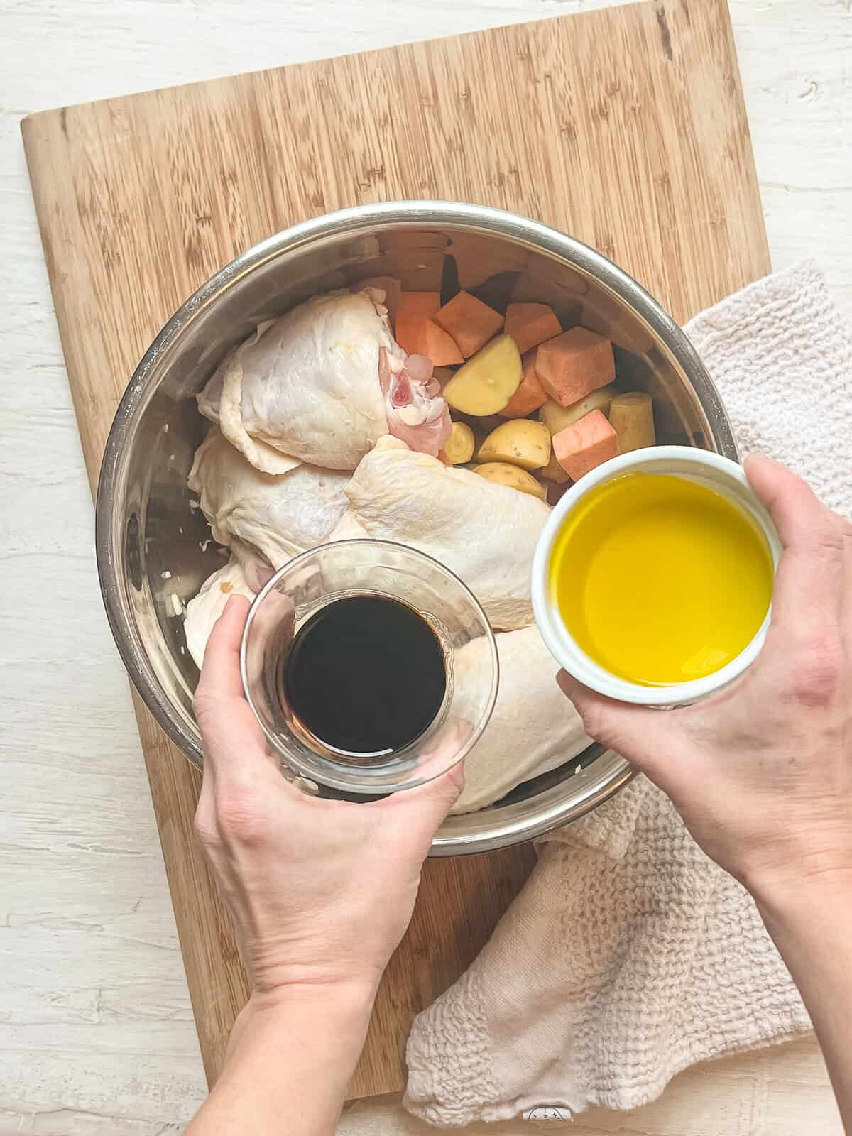 Balsamic vinegar and olive oil being dumped in a bowl with chopped potatoes and carrots and bone-in, skin-on chicken thighs.