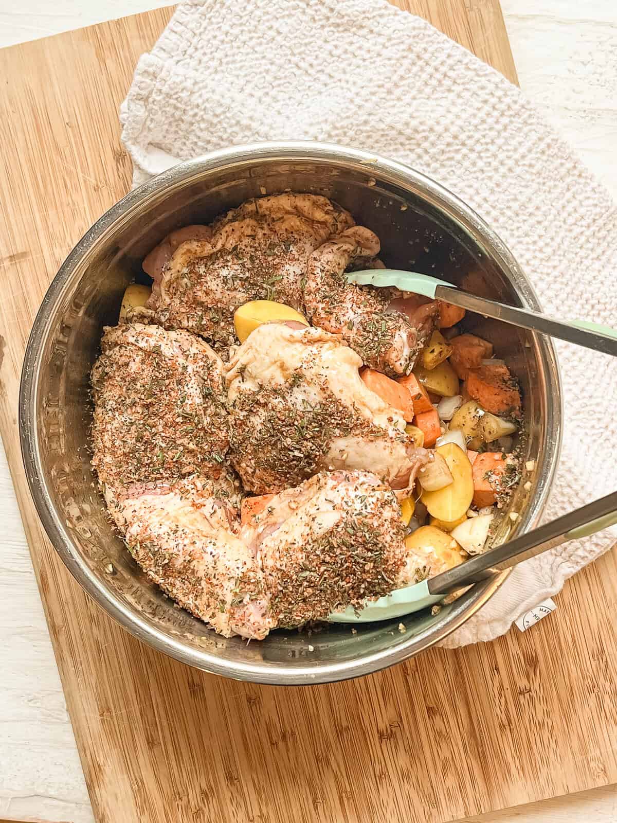 Chopped veggies and raw bone-in, skin-on chicken thighs in a mixing bowl being mixed with dried seasonings.