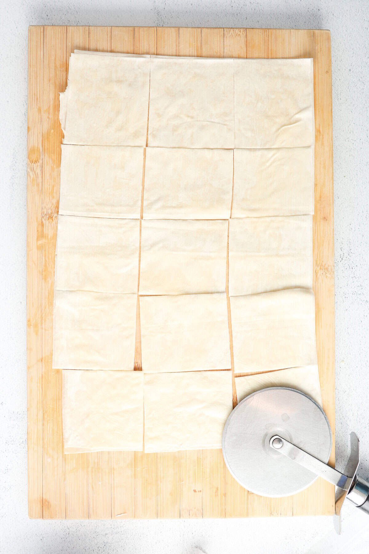 A pizza cutter sitting on a wooden cutting board with a sheet of phyllo dough having been cut into 15 small squares.