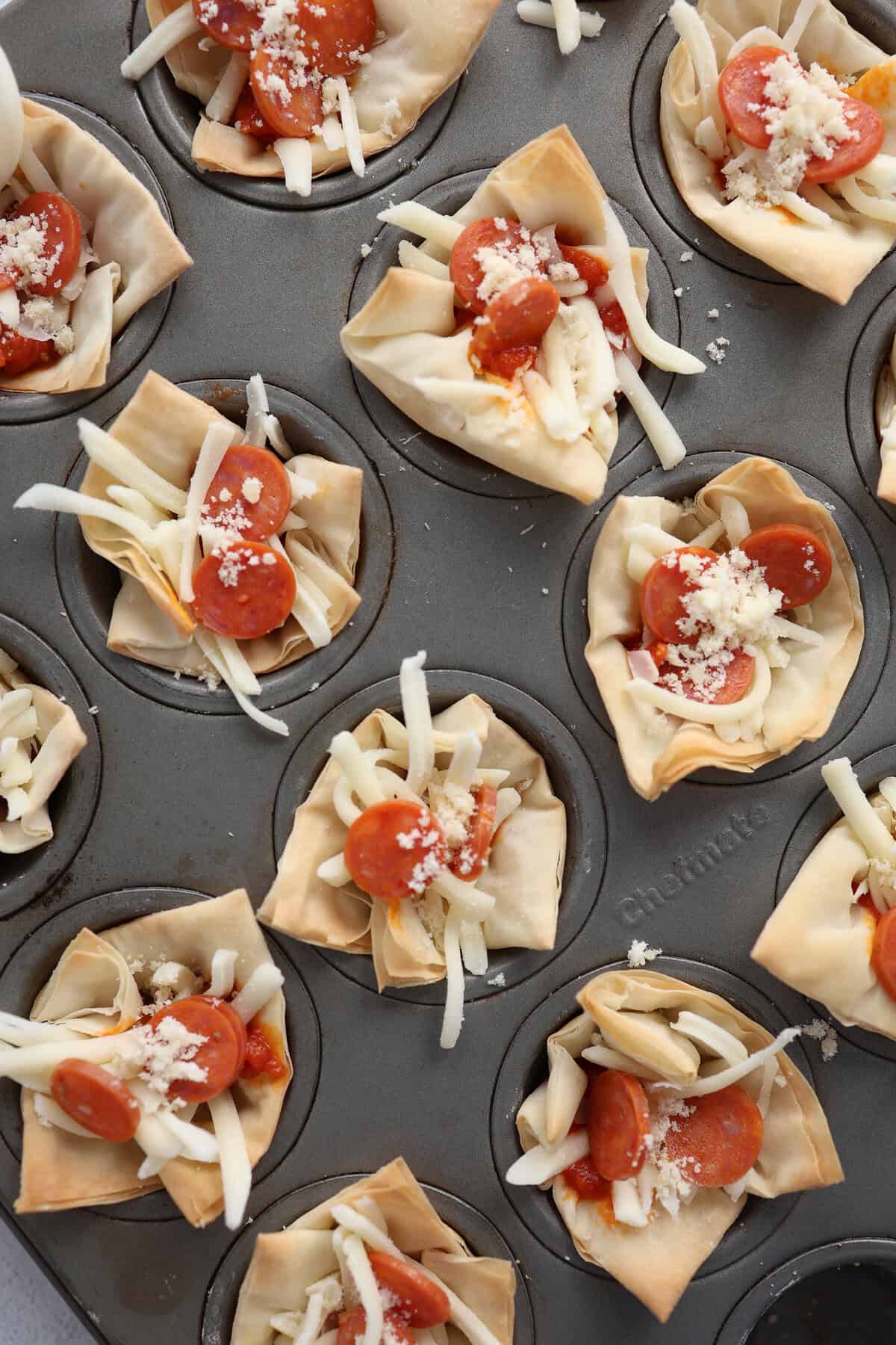 A mini muffin tin with toasted mini phyllo cups, mozzarella cheese, pizza sauce, and mini pepperonis ready to be baked.