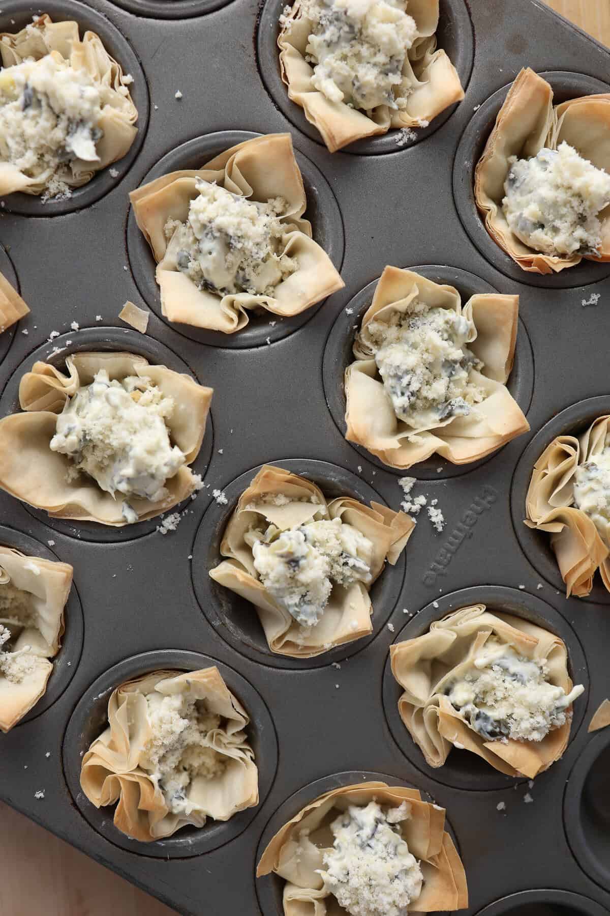 Mini phyllo dough cups in a mini muffin tin with spinach and artichoke dip and grated Parmesan cheese before baking.