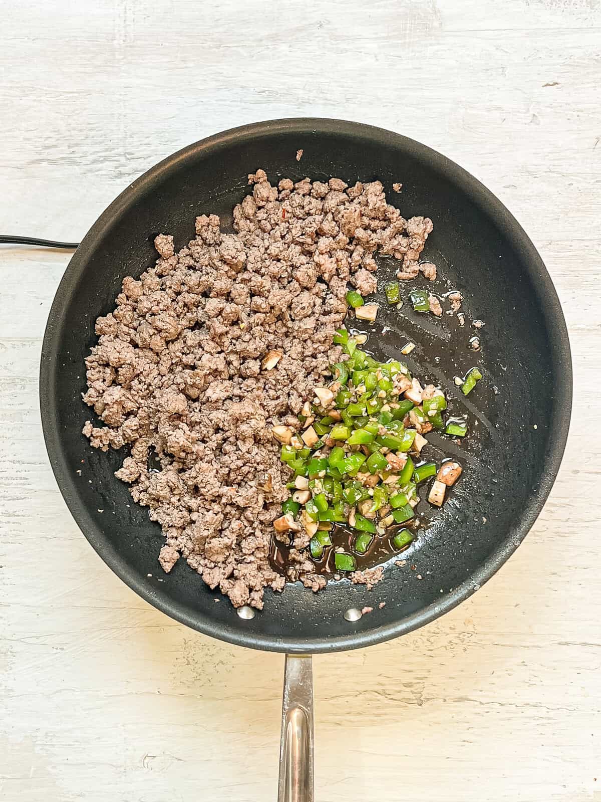 Browned ground beef being mixed with sauteed diced green peppers and mushrooms in a skillet.