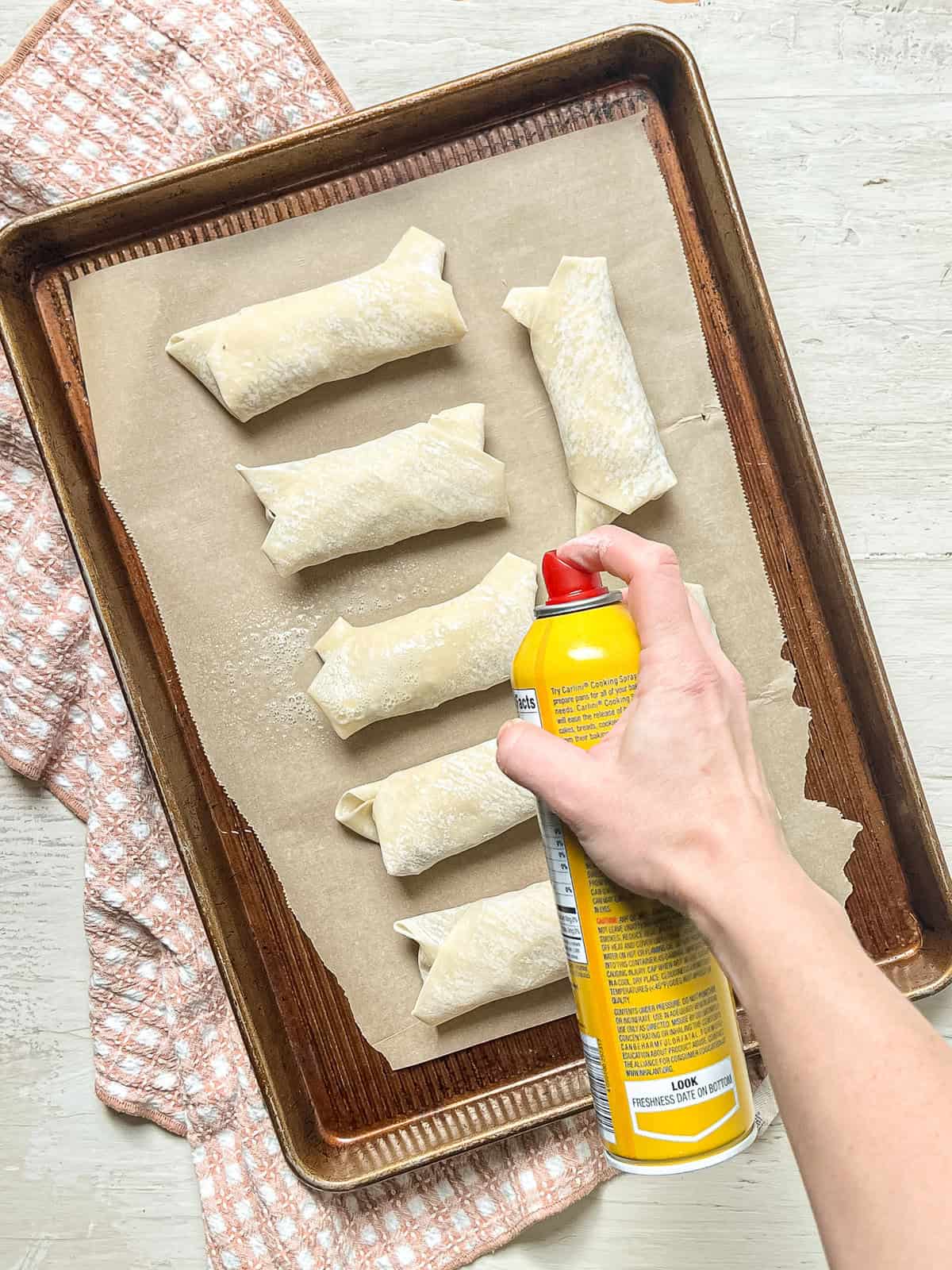 Cheeseburger egg rolls lined up on a parchment-lined baking sheet with someone spraying cooking oil on them.