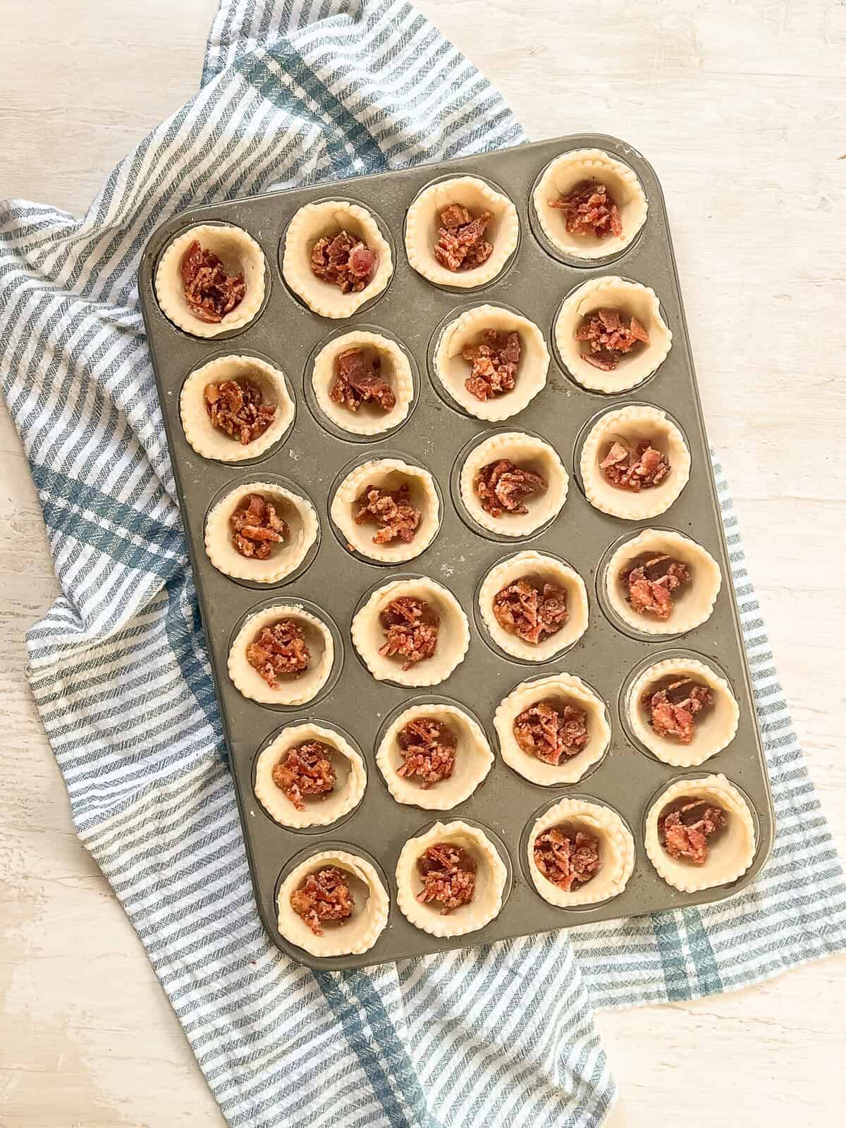 Pie crusts in a mini muffin tin with diced bacon in them.