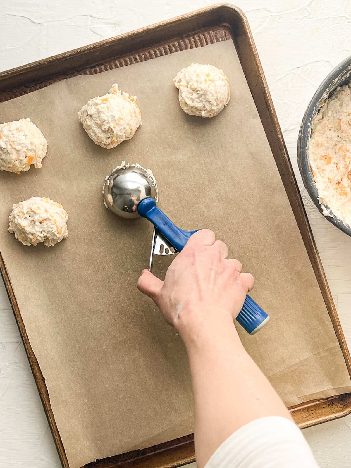 An ice cream scoop putting mounds of drop biscuit dough on a parchment-lined baking sheet.
