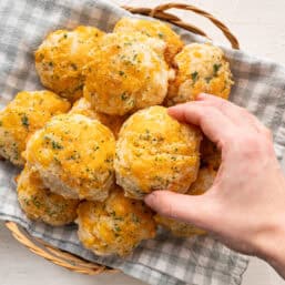 A hand grabbing the top biscuit from a basket of cheddar bay biscuits.
