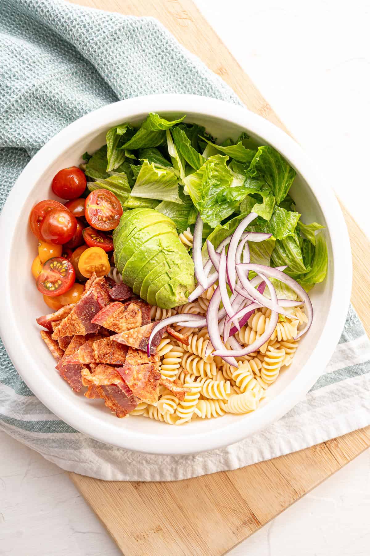 Cooked pasta, chopped lettuce, cherry tomatoes, bacon, and sliced avocado and red onion all in a bowl.