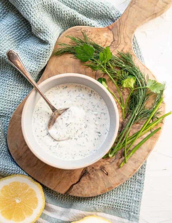 A bowl of homemade ranch dressing on a wooden cutting board with a spoon.