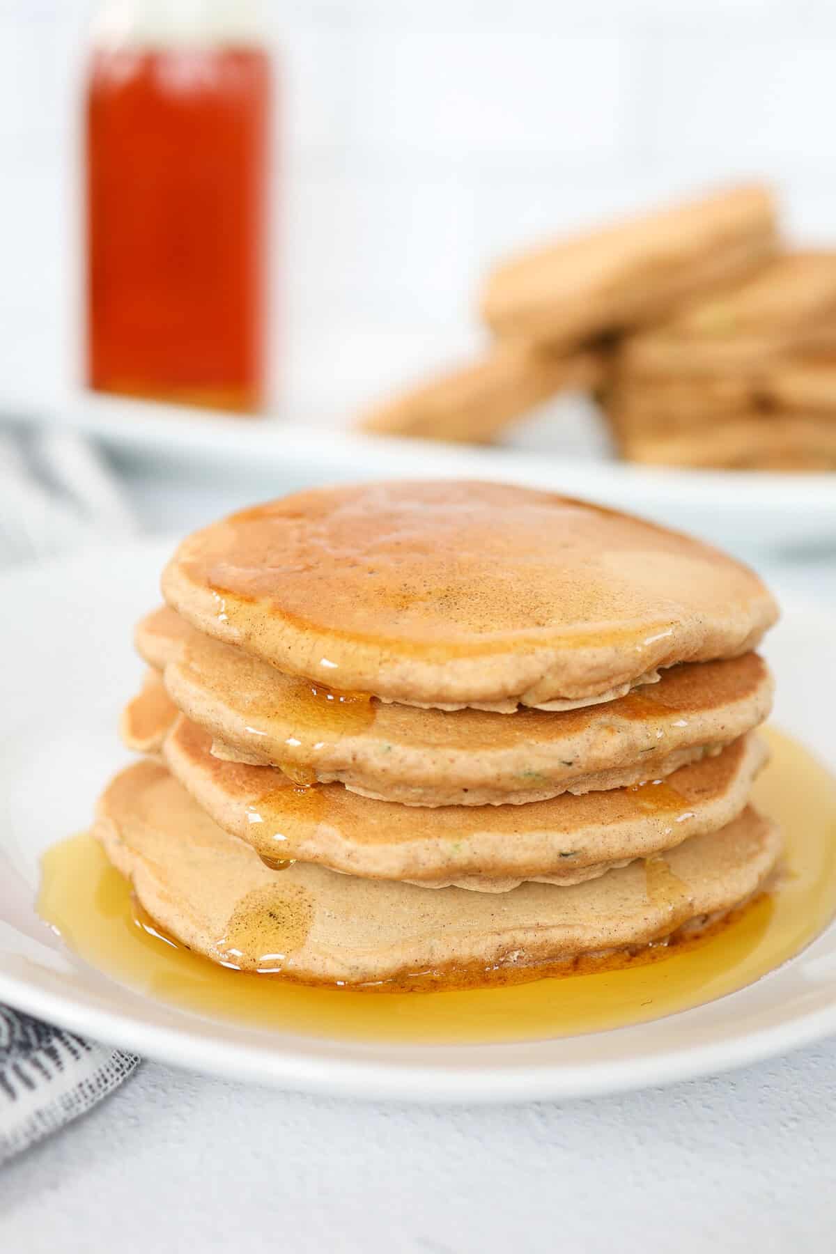 Zucchini pancakes stacked on a plate with maple syrup poured over them.