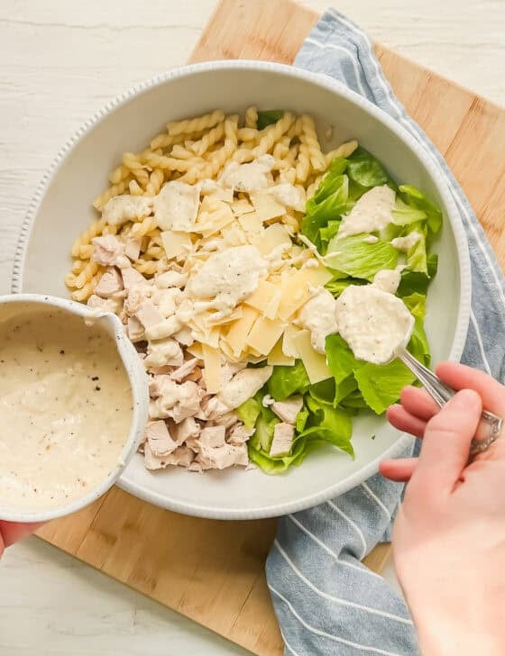 Homemade Caesar dressing being spooned on a bowl with pasta, cooked chicken, chopped Romaine lettuce, and shaved Parmesan.
