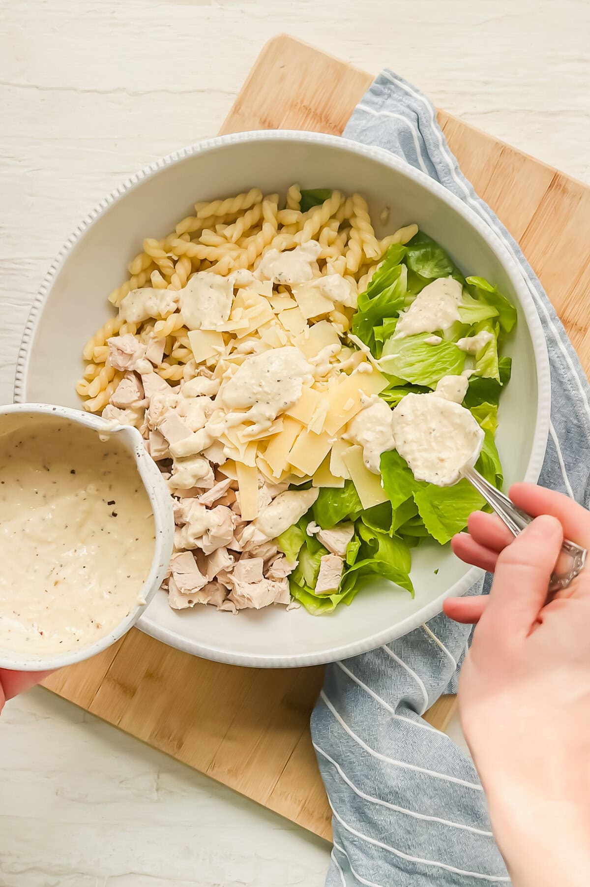 Homemade Caesar dressing being spooned on a bowl with pasta, cooked chicken, chopped Romaine lettuce, and shaved Parmesan.