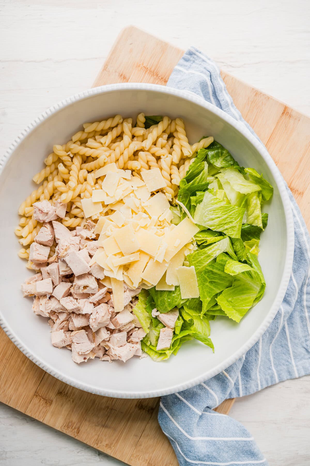 Cooked rotini pasta, chopped Romaine lettuce, cooked cubes of chicken and shaved Parmesan in a bowl ready to be tossed with Caesar dressing.