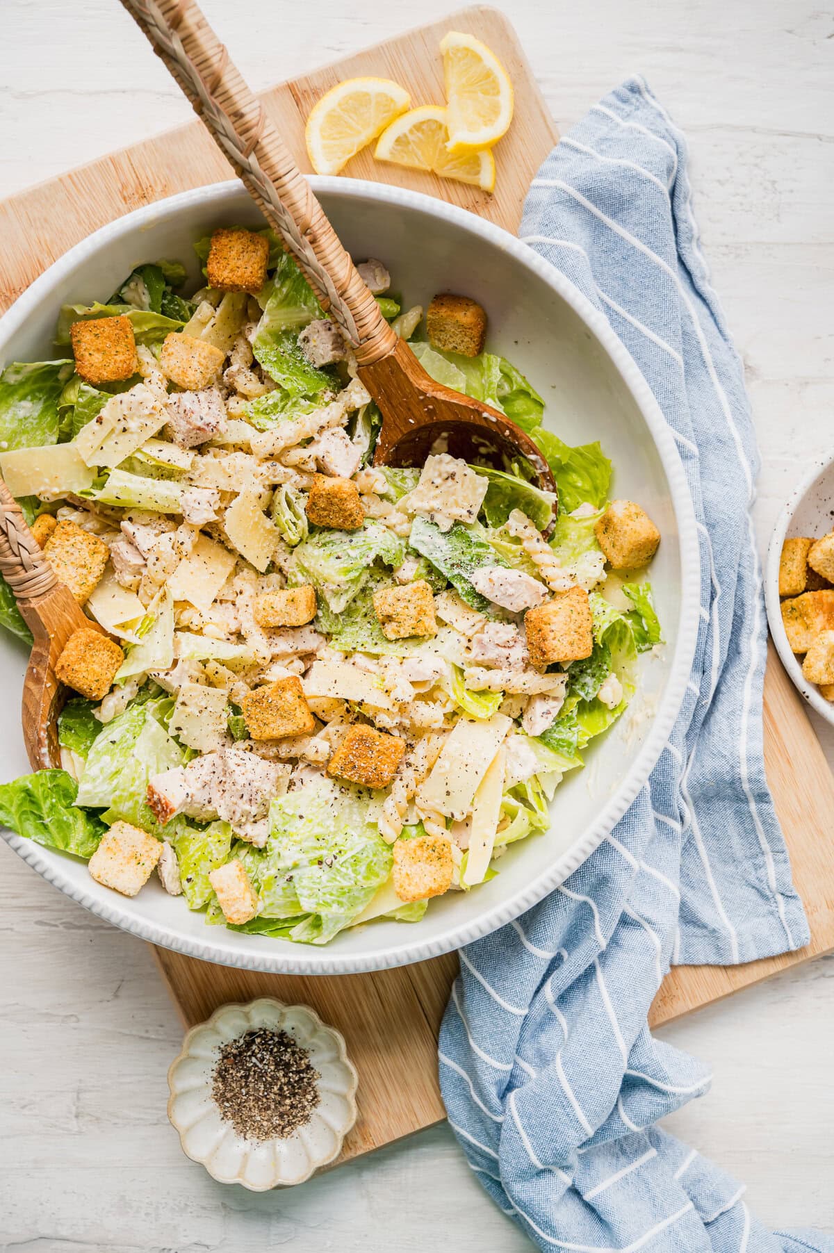 Caesar pasta salad served with two wooden salad tongs and a small dish of ground pepper next to it.