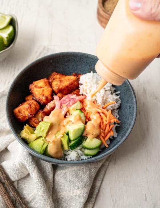 Bang Bang Sauce being squeezed on to a bowl with rice, salmon bites, pickled red onion, avocado, carrots and cucumbers.