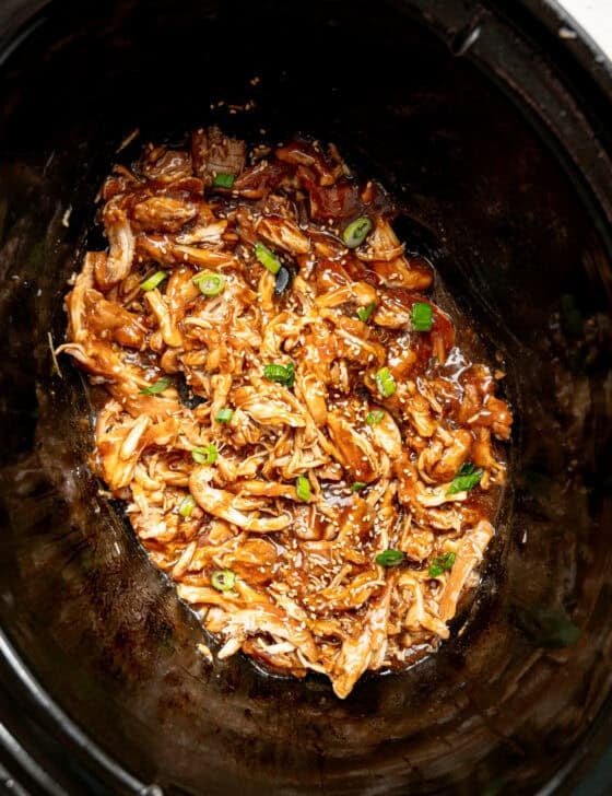 Shredded chicken in a slow cooker covered in teriyaki sauce with sesame seeds and chopped green onions on top.