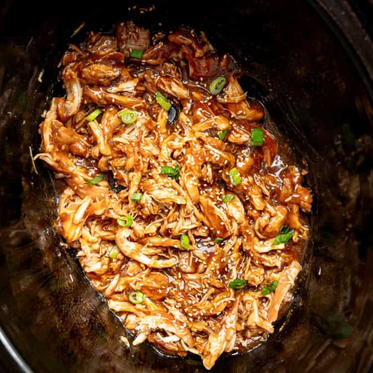 Shredded chicken in a slow cooker covered in teriyaki sauce with sesame seeds and chopped green onions on top.
