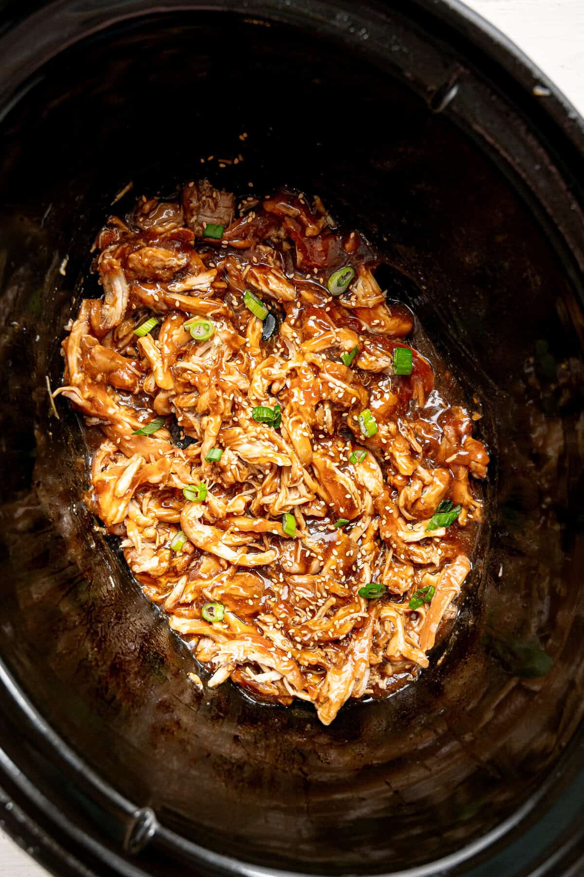 Shredded chicken in a slow cooker covered in teriyaki sauce with sesame seeds and chopped green onions on top.