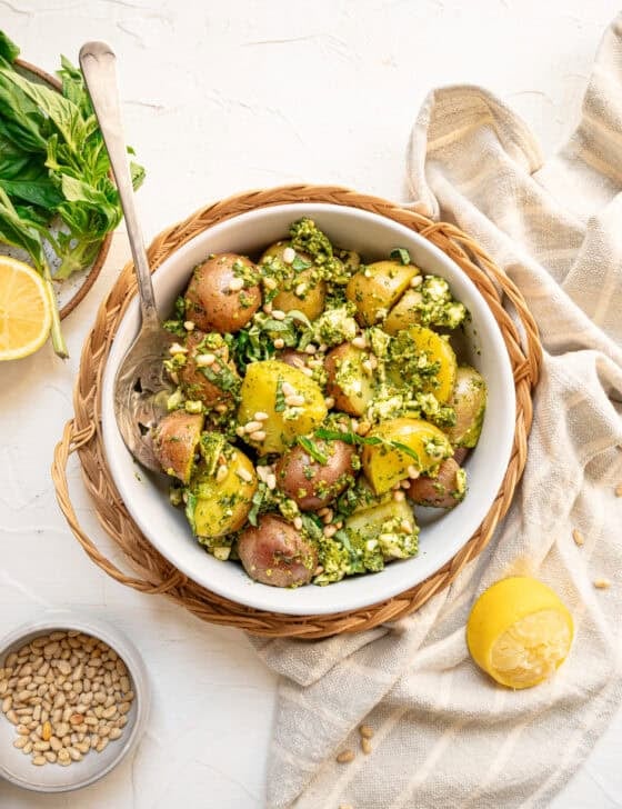 Pesto potato salad in a white bowl with fresh basil and pine nuts on top.
