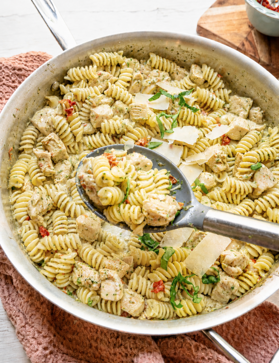 A serving spoon lifting creamy chicken pesto pasta from the skillet.