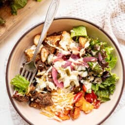 A cheeseburger bowl made up of roasted potato bites, crumbled ground beef, shredded cheese, lettuce, tomatoes, and pickled onions.